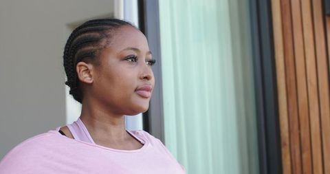 Contemplative african american woman standing by sliding glass door wearing pastel pink top