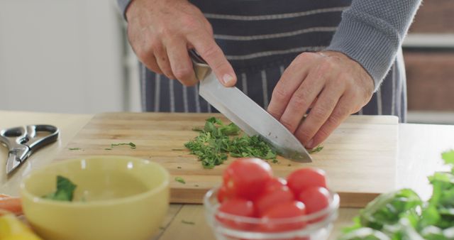 Man chopping fresh green herbs on wooden cutting board in kitchen. Visible tomatoes and other ingredients suggest this is part of preparing a meal. Useful for content related to cooking, home kitchens, food preparation, and culinary arts.