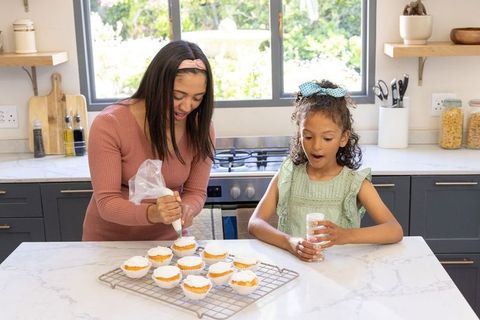 Diverse mother and daughter decorating cupcakes on countertop in sunlit home kitchen