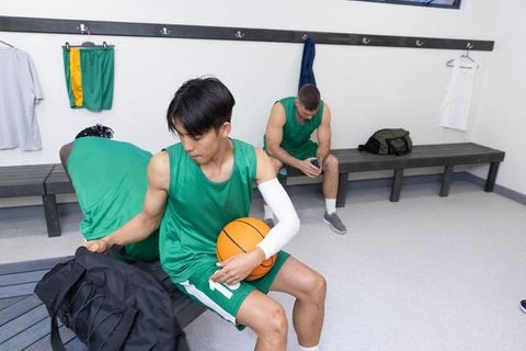 Diverse basketball team sitting in locker room holding basketball and reaching into duffel