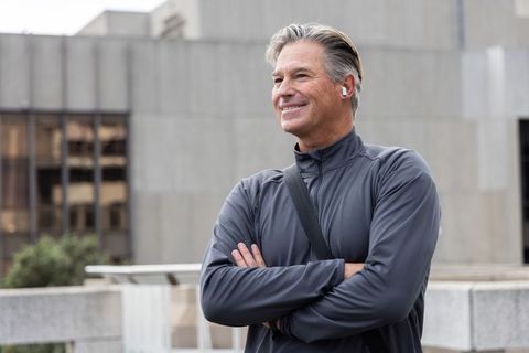Confident mature man standing on urban terrace wearing wireless earbuds and crossbody bag