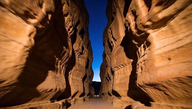 Sunlit narrow sandstone slot canyon leading along paved path toward bright blue sky