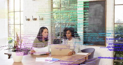 Collaborating women reviewing laptop and documents in modern office with code overlay