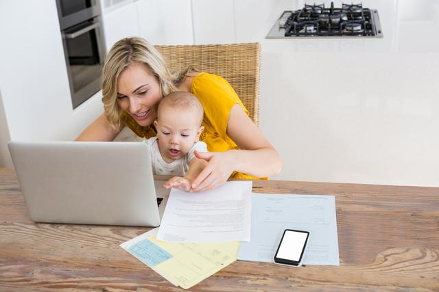 Mother using laptop with baby boy in kitchen at home
