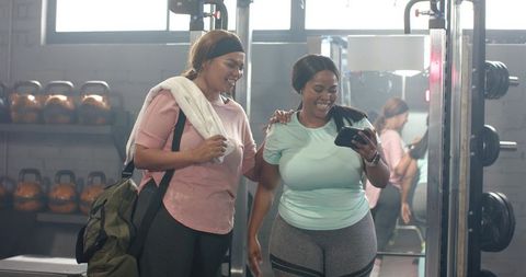 Diverse women checking smartphone after workout, smiling together in gym weightroom