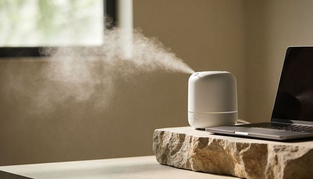 Minimal white ultrasonic diffuser releasing fine mist on natural stone desk beside laptop