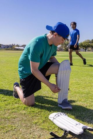 Teen fastening cricket leg pad on grassy field while teammate walks by during practice