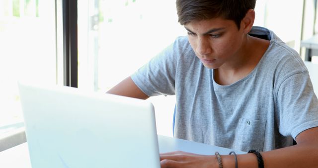 Teenager boy concentrating while using laptop in light-filled room. Ideal for educational materials, technology-focused campaigns, modern learning environments, student life articles, and youth culture concepts.