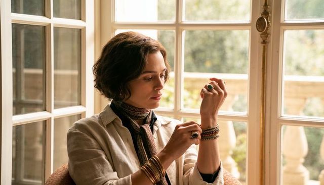 Woman adjusting layered bracelets and rings by sunlit casement window, calm contemplation