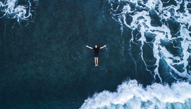 Aerial man floating face up in deep teal ocean amid rolling breaking waves and white foam
