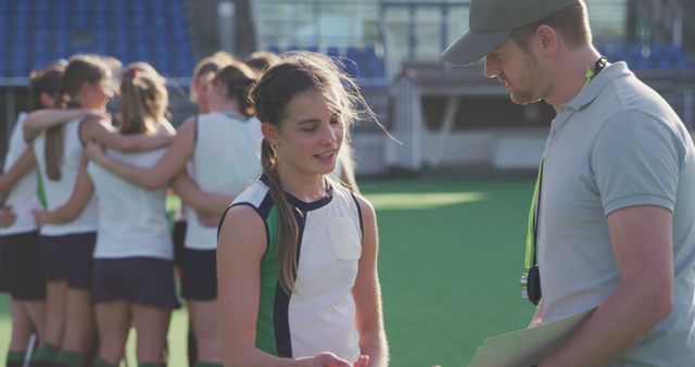 A young field hockey player listens intently to her coach's feedback during practice on the hockey field. Teammates in the background chat and support one another, emphasizing teamwork and camaraderie. This image is ideal for depicting youth sports training, coaching, physical education programs, and promoting teamwork and mentorship in sports.