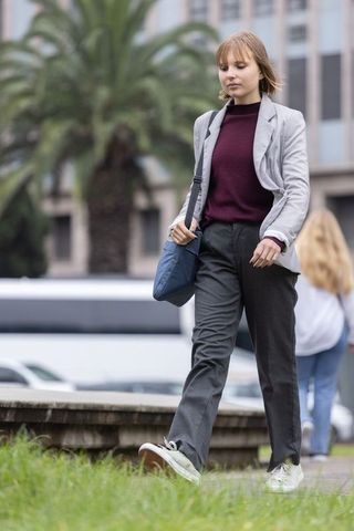 Young woman walking along grassy fountain carrying blue crossbody bag wearing gray blazer