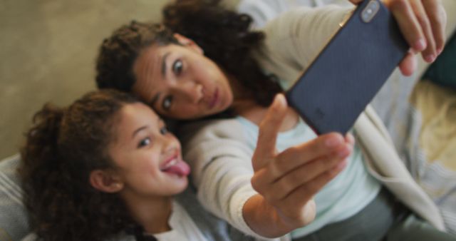 Mother and daughter lying on a couch, having fun taking a playful selfie. Both are making silly faces, showcasing their bond in a lighthearted moment. Ideal for illustrating family joy, parent-child relationships, and candid home scenarios. Suitable for use in family-oriented promotions, parenting blogs, and technology advertisements.