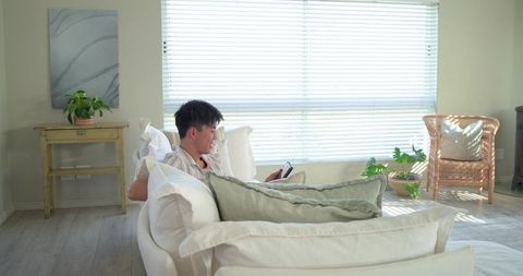 Asian man reclining on light sofa holding smartphone among cushions in sunlit living room