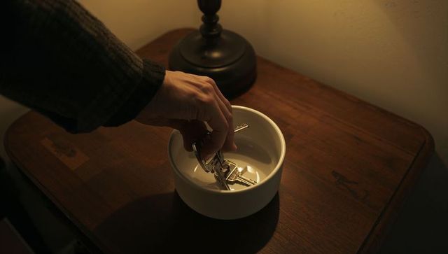 Hand placing keys in ceramic bowl on wooden nightstand under warm lamp light