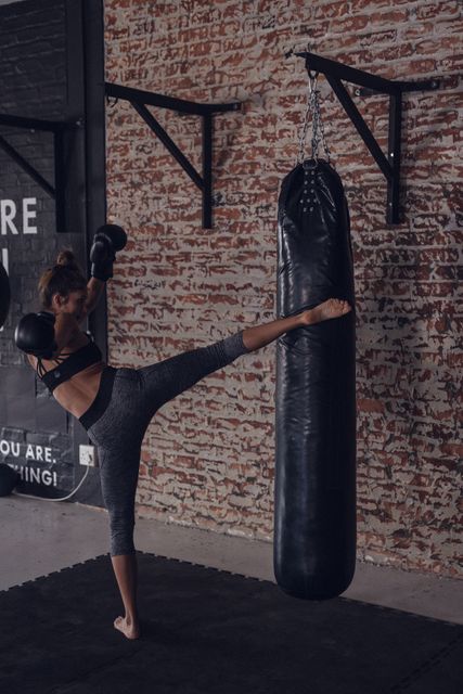 This scene shows a dedicated young female boxer practicing kicks at a gym. The setting features a rustic brick wall, adding an industrial aesthetic. Useful for illustrating concepts related to women's fitness, strength training, boxing practice, and athletic dedication.