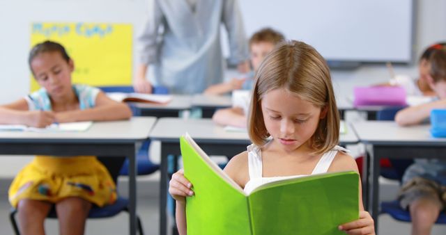 This image depicts a young girl intently reading a green book. Other students appear engaged in independent activities in the background with a teacher overseeing. This picture can be used for educational articles, school-related advertisements, or websites focused on primary education. Perfect for themes on studying, focus, and academic environments.