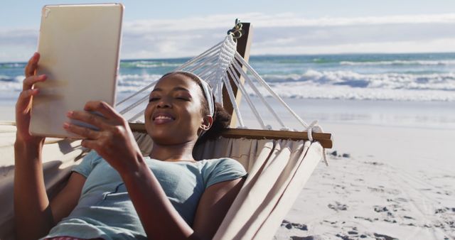 Woman smiling while using tablet and relaxing in hammock at beach. Excellent for promoting travel destinations, beach resorts, vacation plans, leisure activities, digital nomad lifestyle, and technology integration in everyday life.
