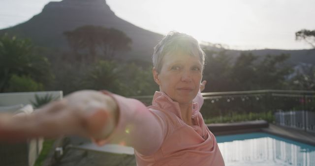 Confident senior woman extends arm while posing outdoors against mountain background with clear poolside visible. Ideal for use in promoting healthy lifestyle, senior fitness, aging gracefully, outdoor retreats, vacation destinations, and wellness center advertisements.
