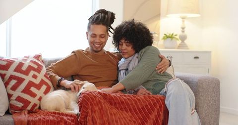 African american couple cuddling on sofa petting puppy under cozy rust-red blanket at home