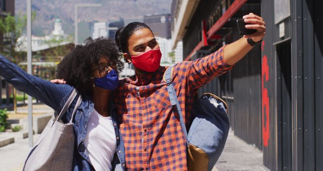 Diverse couple wearing face masks embracing and taking a selfie. digital nomads on the go during coronavirus covid 19 pandemic.