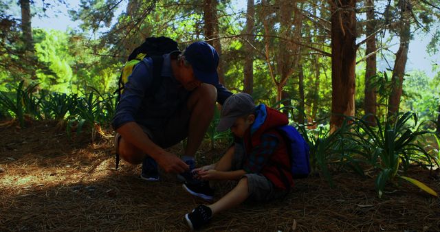 Father helping his young son tie his shoes in a forest setting during a hike. Scene emphasizing family bonding, teaching moments, and outdoor activities. Perfect for themes related to parenting, family adventures, nature hikes, and father-son relationships.