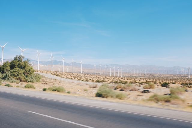 Photo of sizable wind farm in desert landscape with clear blue sky. Wind turbines lined in rows stretch into distance, highlighting advancement in renewable energy and sustainability. Ideal for topics related to renewable energy, environmental conservation, sustainable technology, and clean power initiatives. Scenic nature view and clear skies evoke serenity.