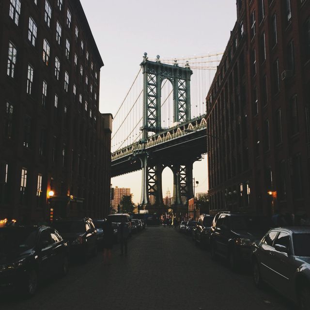Manhattan Bridge viewed through a street in DUMBO, Brooklyn, creating a striking urban scene at sunset. Buildings and parked cars frame the iconic structure, highlighting the blend of old and new architecture. Ideal for travel blogs, city-related articles, or promotional material for tourism in NYC.