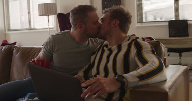 Happy caucasian gay male couple sitting on couch with cups, using laptop and kissing in living room. Togetherness, relationship, communication and domestic life, romance, unaltered.