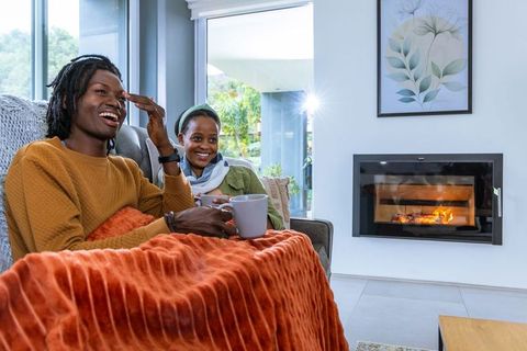 African american couple relaxing and laughing with mugs on sofa by modern fireplace with warm throw