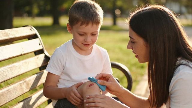 Mother caring for young boy's scraped knee on park bench using blue cloth in sunlight