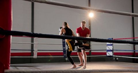 Trainer holding kick pad while female fighter delivering kick in boxing ring with dramatic flare