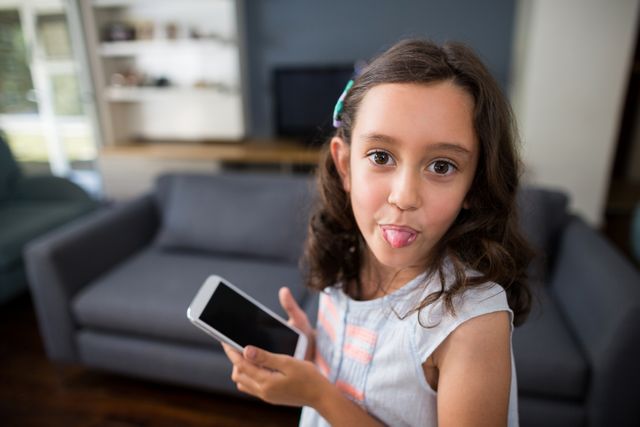 Girl sticking out tongue while holding mobile phone in living room