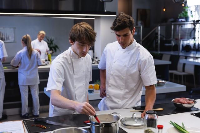 Two biracial male chefs cooking in a modern busy kitchen, mixing contents in a pan on hob. Cookery class at a restaurant kitchen. Workshop cooking food.