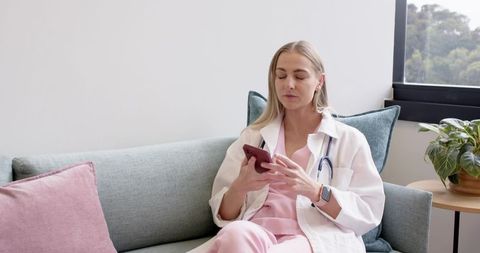 Nurse relaxing on sofa using smartphone in pastel healthcare lounge, stethoscope