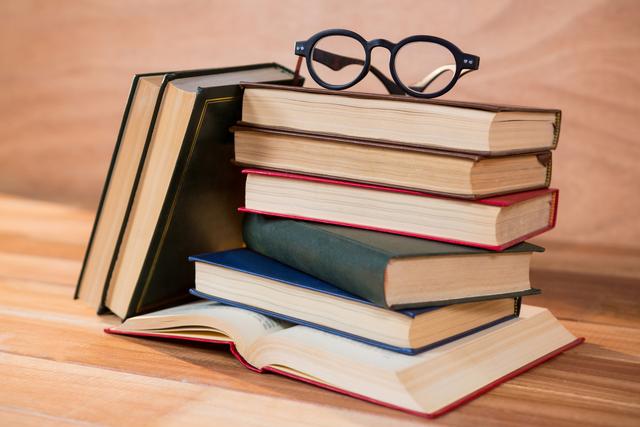 This image shows a stack of books with a pair of glasses resting on top, placed on a wooden table. An open book is also visible. This image can be used for educational content, library promotions, book club advertisements, or articles related to reading and literature.
