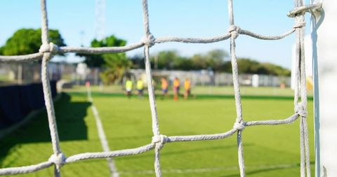 Close-up white knotted soccer goal net with blurred players wearing bright bibs on grass pitch