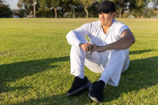 Athlete sitting on sunlit field wearing all-white kit and black cap, casting long shadow