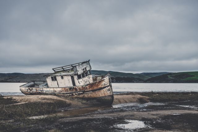 A rusty, abandoned fishing boat sits on a muddy shoreline, under a grey, cloudy sky. The scene creates a moody and atmospheric feeling. Perfect for use in editorials about coastal decay, maritime history, the passage of time, or environmental themes. Alternatively useful as a compelling visual for websites, blogs, or articles discussing derelict vessels and forgotten places.