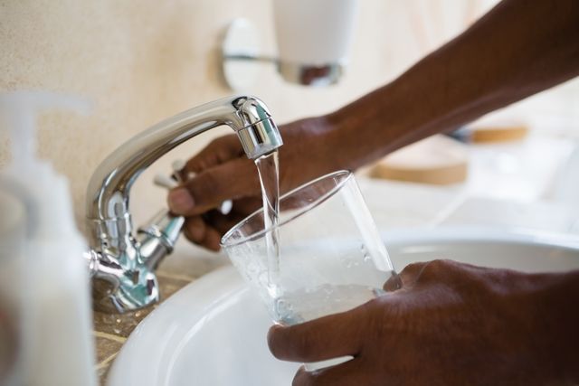 This image shows a man filling a glass with water from a bathroom sink faucet. It can be used to illustrate concepts of hygiene, daily routines, hydration, and home life. Suitable for articles, blogs, and advertisements related to water conservation, health, and wellness.