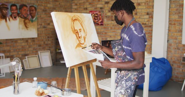African american male painter in face mask painting from photograph on canvas in artist studio. art, creative and leisure time during covid 19 pandemic concept.