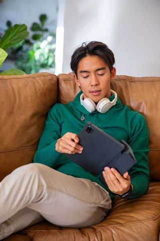Asian man relaxing on tan couch using tablet with white headphones in sunlit living room