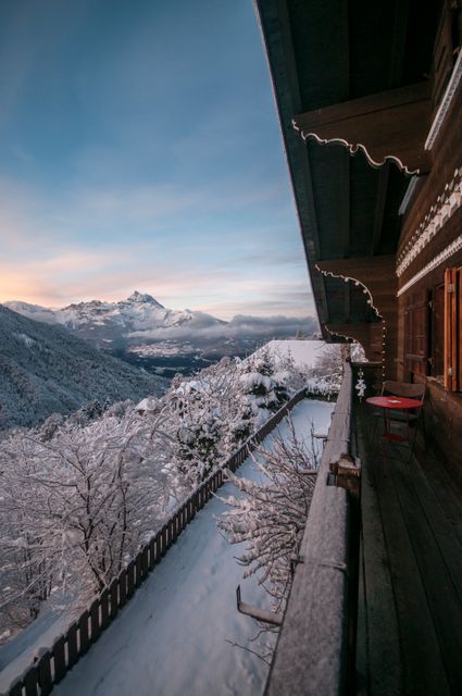 Wooden balcony of a chalet overlooking a snowy mountain landscape at sunrise. Snow-covered trees and mountains create a serene and tranquil atmosphere. Ideal for illustrating winter travel destinations, mountain holidays, idyllic retreats, or scenic outdoor environments.