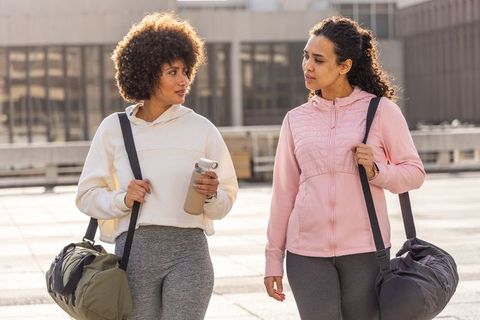 Two friends walking urban plaza in athleisure carrying gym bags and water bottle