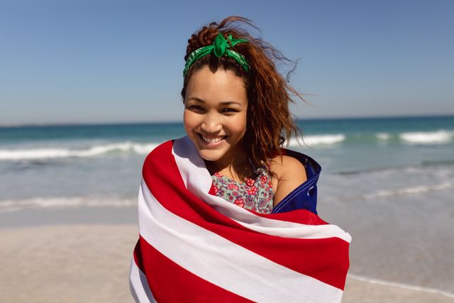 Biracial woman wrapped in towel, standing on beach, smiling. Ideal for promoting summer vacations, beach activities, travel destinations, and leisure time. Perfect for advertisements, travel blogs, and lifestyle magazines.