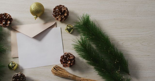Christmas holiday card mockup featuring a blank card with an envelope on a wooden table. Surrounding items include green branches, pine cones, gold baubles, and small bells. Ideal for holiday greeting card designs, festive invitations, and seasonal promotional materials.