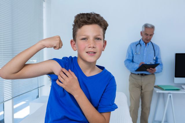 Portrait of boy flexing his biceps in the clinic and doctor standing behind him