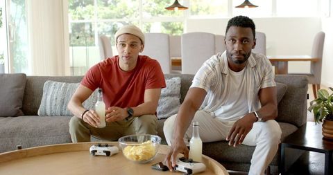 Diverse male friends playing video games and sharing chips and drinks on modern couch