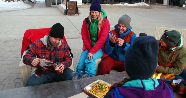 A group of friends enjoying a meal and drinks outdoors after participating in winter activities. Everyone is dressed in warm clothing, sitting casually and laughing, demonstrating camaraderie and the enjoyment of shared experiences. Perfect for concepts related to friendship, outdoor activities, winter fun, and social gatherings.