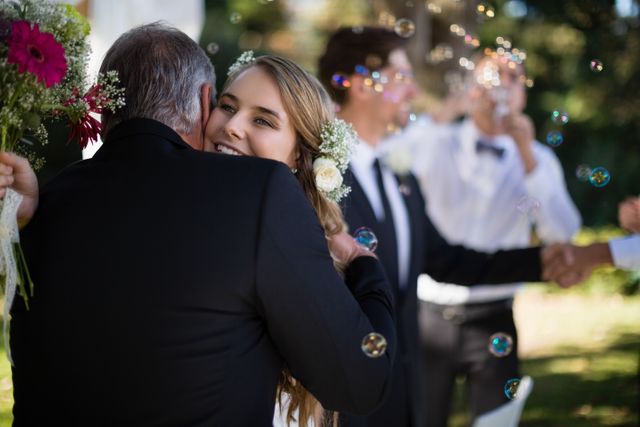 Father embracing his daughter during an outdoor wedding ceremony. The scene captures a heartfelt moment filled with love and joy, with bubbles floating around and other guests in the background. Ideal for use in wedding planning materials, family celebration promotions, and articles about special family moments.